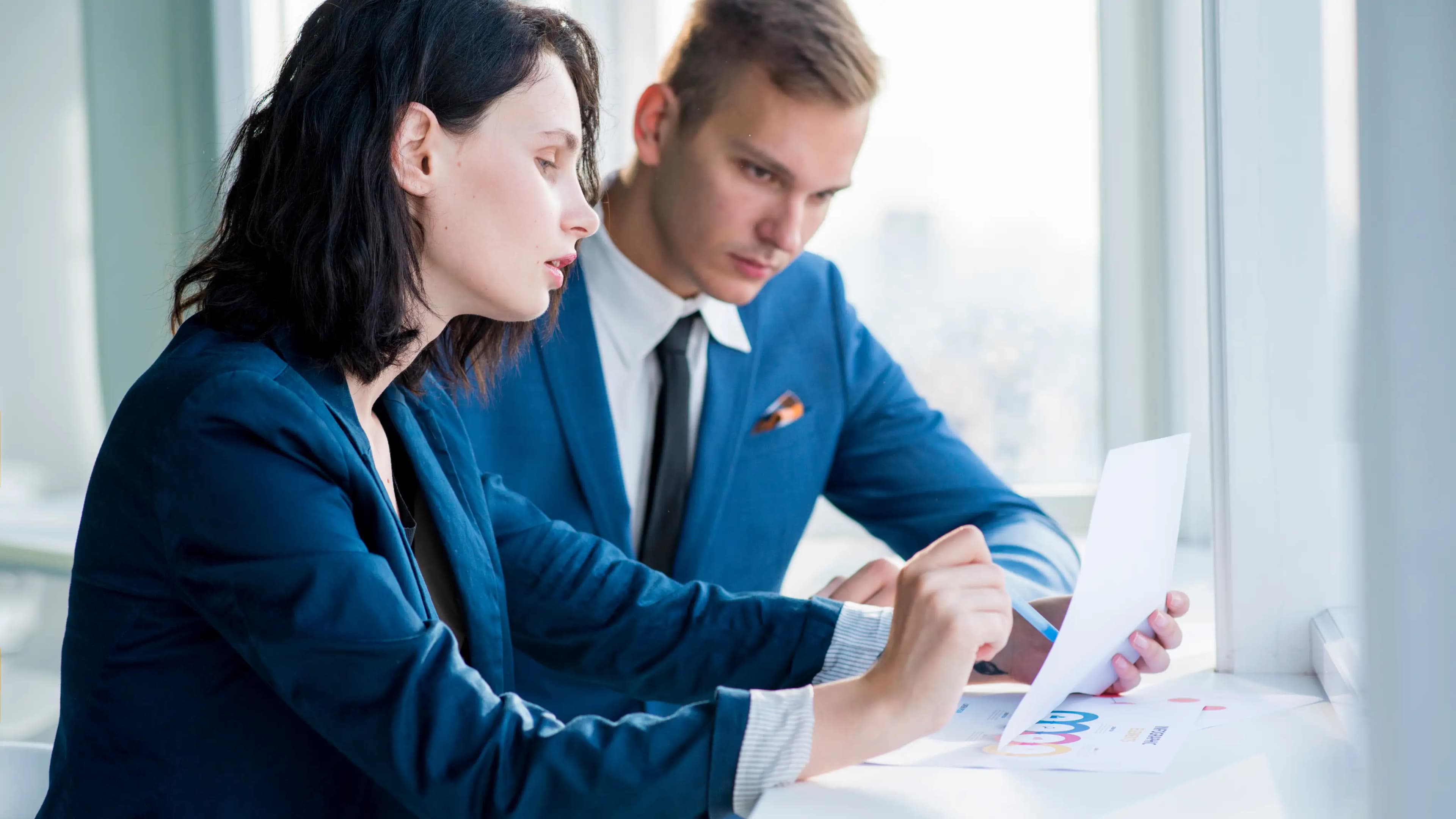 Two business professionals in formal attire sitting at a desk by the window, one holding and pointing to a document with graphs, demonstrating collaborative and accurate EIN registration.