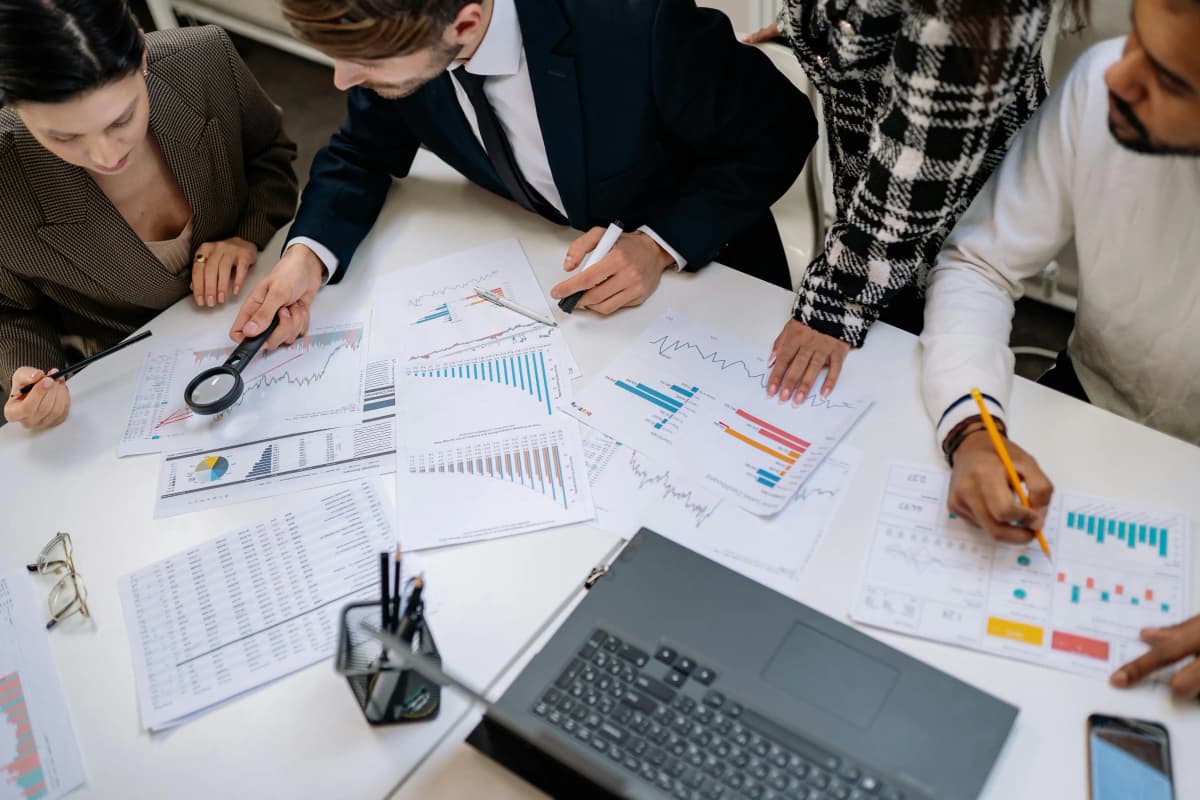 Four business professionals gathered around a desk reviewing charts and paperwork, analyzing data and compliance requirements, illustrating expert teamwork in obtaining business licenses.