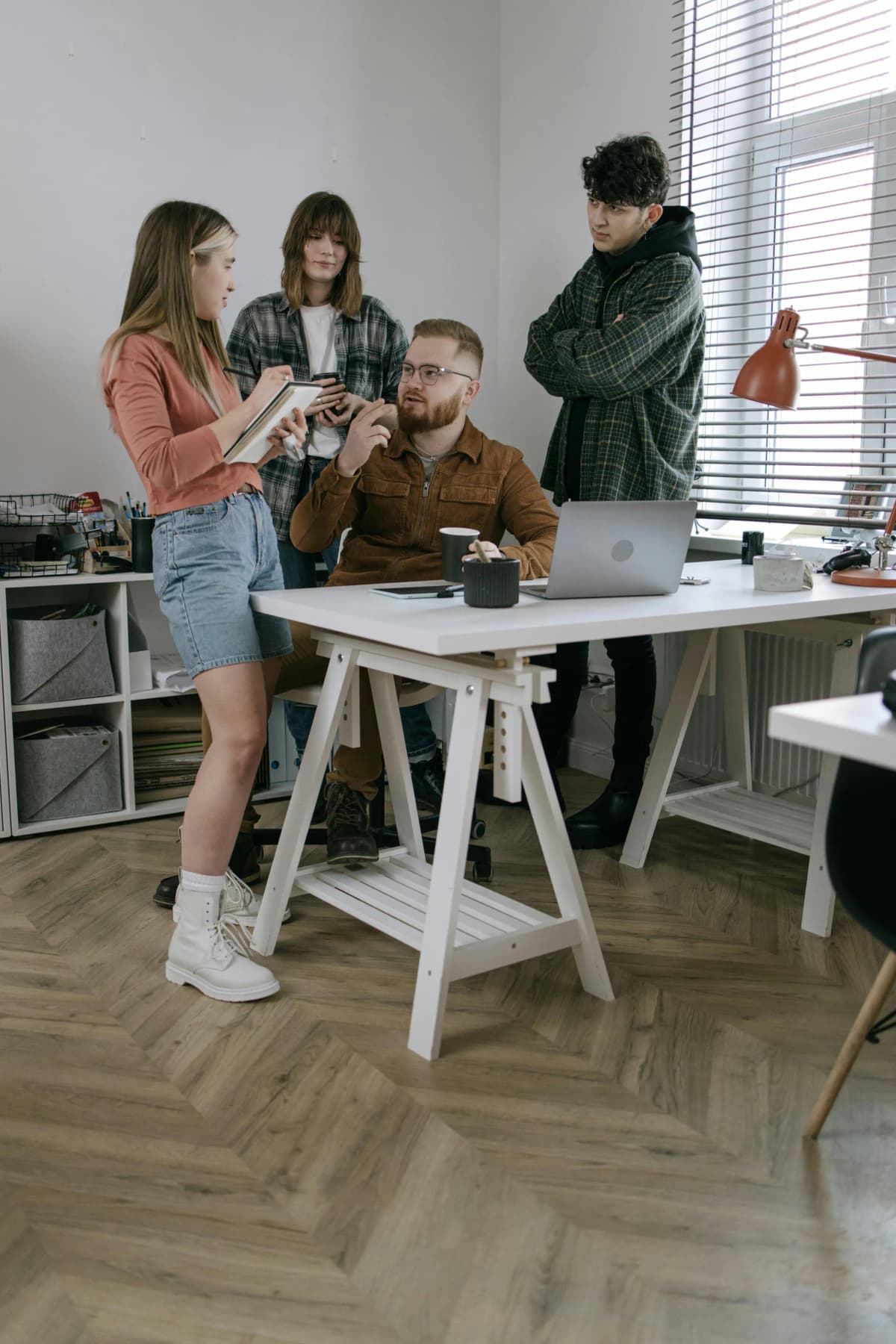 Four business professionals in a modern office, three standing and one seated at a desk with a laptop and lamp, one taking notes in a notebook while another shares ideas and the group listens, illustrating collaborative planning and teamwork for trademark registration and brand protection.