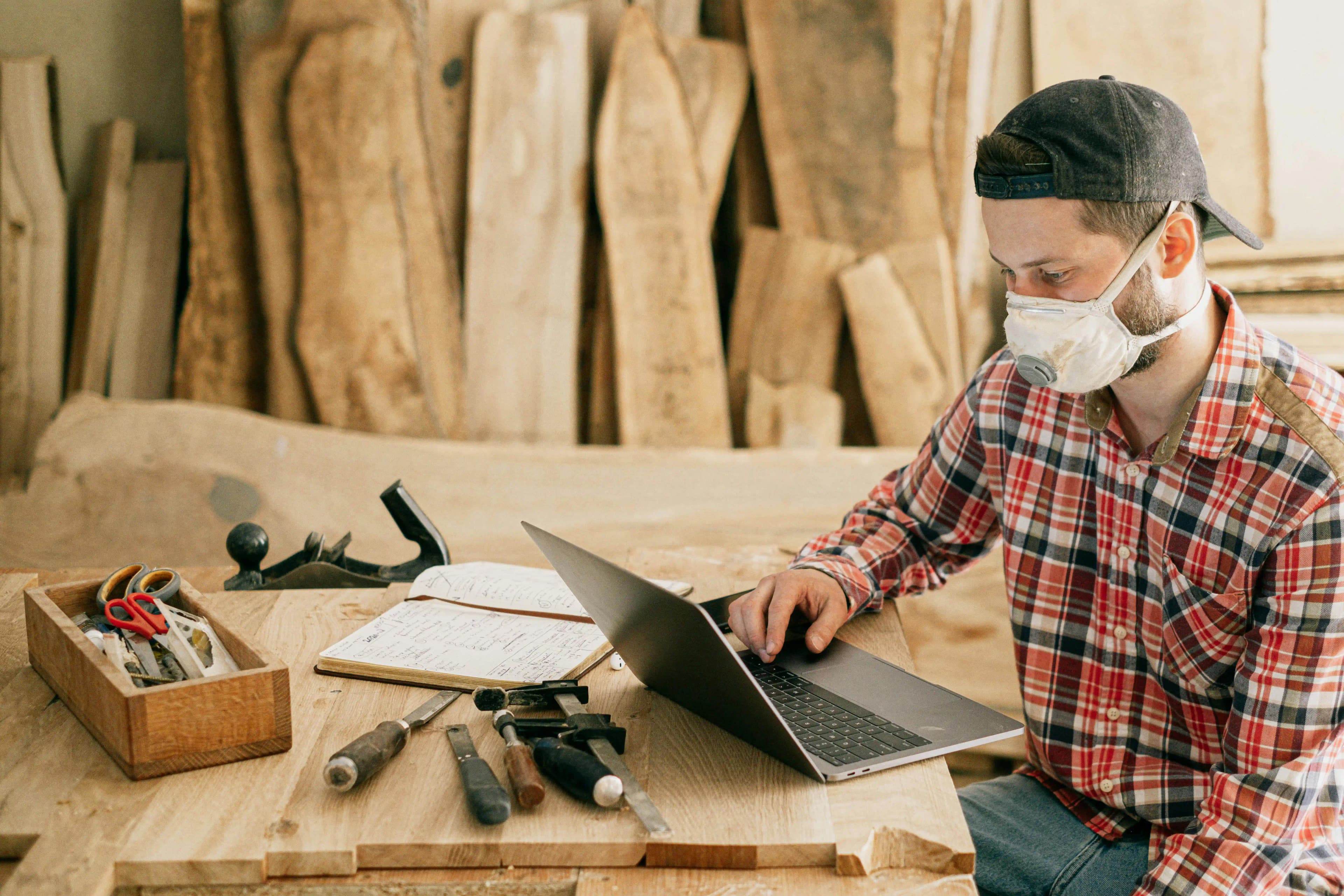 Woodworker sitting at a workbench with tools and a laptop, reviewing digital documents for year-end annual reporting.